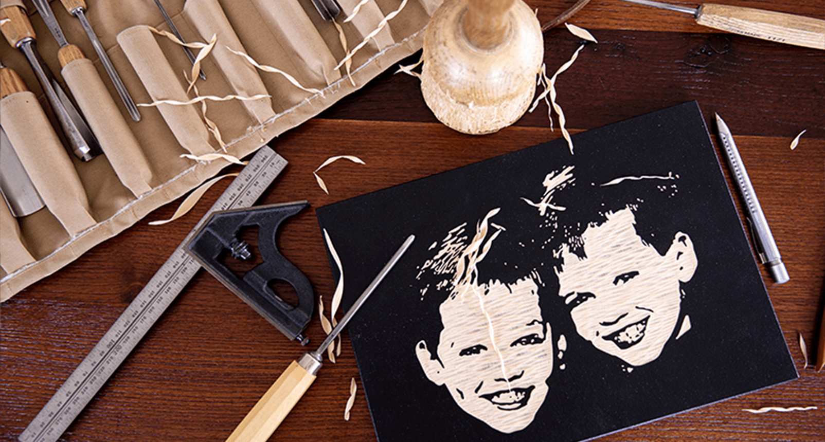 Ein geschnitztes Porträt von zwei lächelnden Gesichtern auf schwarzem Papier mit Schnitzwerkzeugen und Holzspänen.